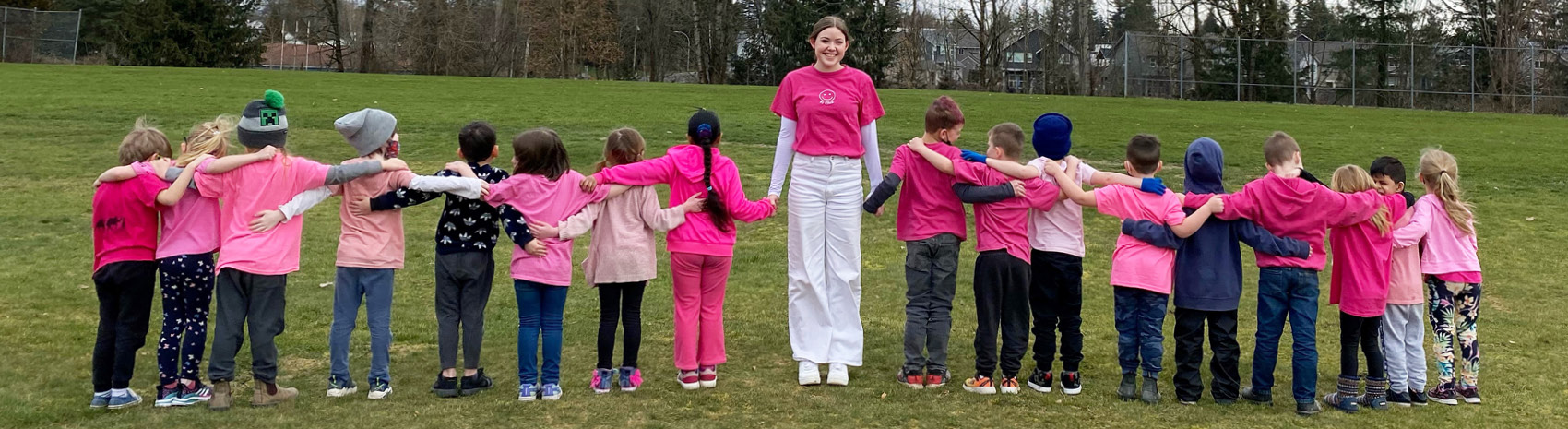 A teacher candidate in a row of young students, all holding hands and wearing pink shirts.